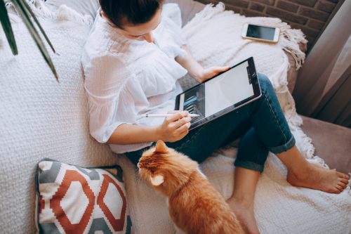 woman-sat-on-a-sofa-working-on-a-tablet-with-her-ginger-cat