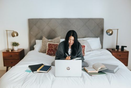 a-woman-sitting-on-a-bed-in-front-of-a-laptop-writing-in-a-notebook