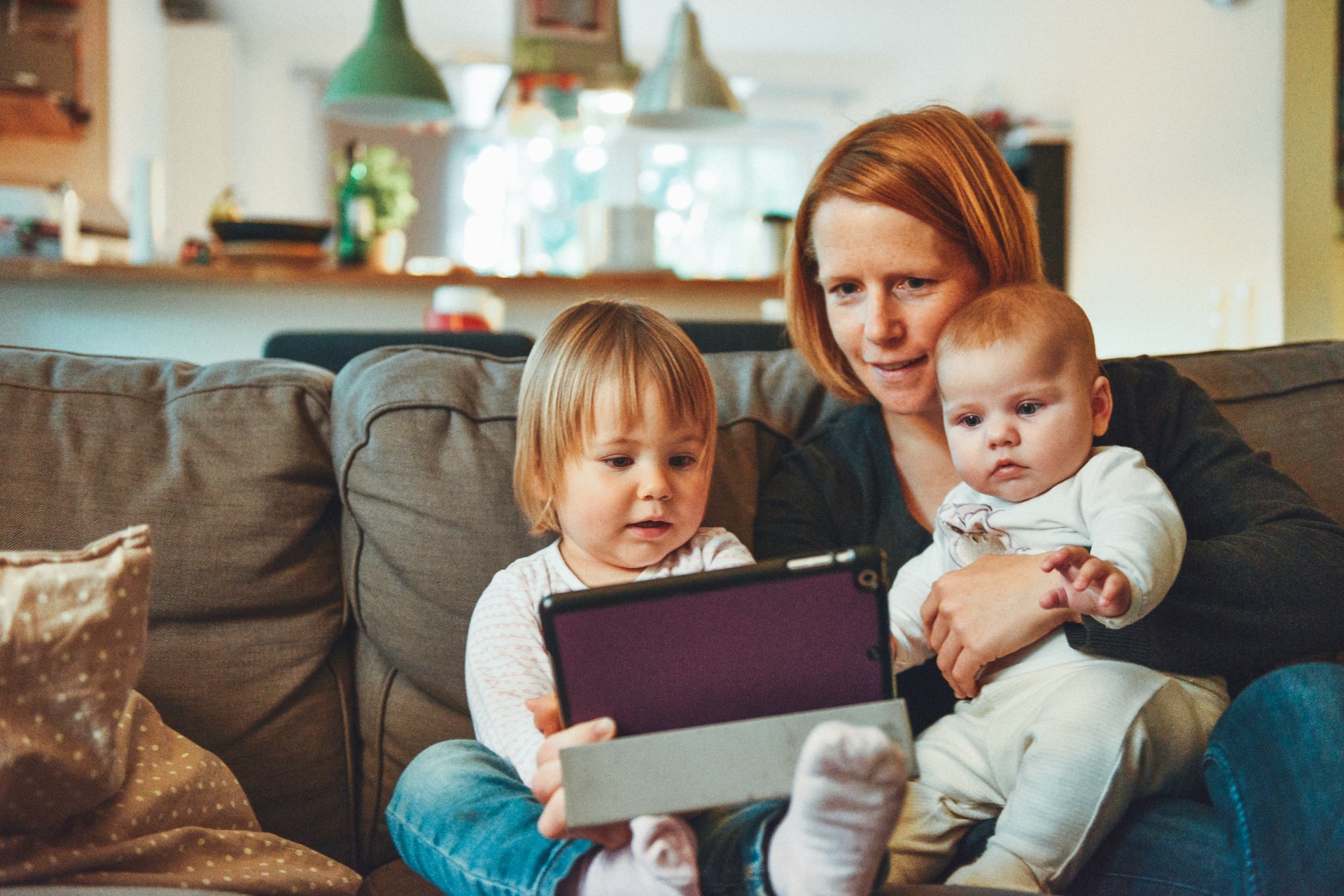 two-babies-and-mother-sitting-on-the-sofa-holding-one-baby-and-looking-at-a-tablet