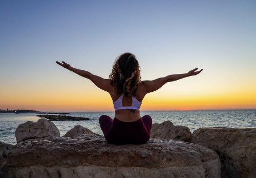 woman-sitting-on-a-rock-next-to-the-sea-with-her-arms-out-wide-and-back-to-the-camera