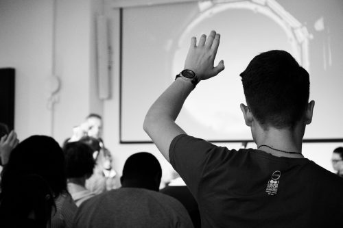 black-and-white-image-of-a-person-from-behind-putting-their-hand-up-to-answer-a-question